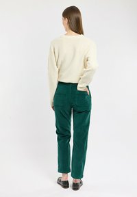Pull en tricot crème avec des manches texturées associé à un pantalon en velours côtelé vert foncé avec deux poches arrière et une coupe décontractée. Chaussures noires à enfiler.