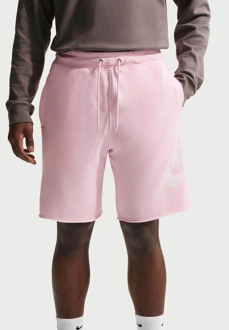 Vaaleanpunaiset puuvillahousut, joissa on joustava vyötärönauha, kiristysnyöri, sivutaskut ja suuri valkoinen Nike-logo vasemmassa lahkeessa.