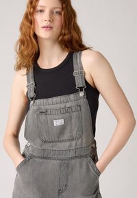 Gray denim overalls feature adjustable straps, front pocket, side buttons, and relaxed fit. Worn over a black ribbed tank top.