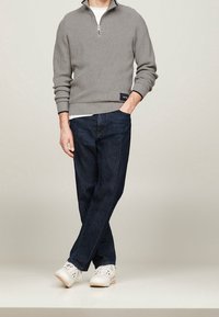 Mann, der mit gekreuzten Beinen steht, trägt einen grauen Zipfeljacke, dunkelblaue Jeans, weiße Sneaker und ein weißes Unterhemd vor einem schlichten Hintergrund.