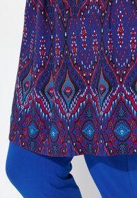 Leicht abgerundete Tunika mit einem mehrfarbigen geometrischen Muster in Lila, Rot und Türkis auf einem dunklen Hintergrund, kombiniert mit blauen Leggings.