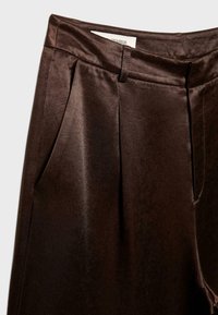 Gros plan sur un pantalon sur mesure brun foncé brillant avec poches latérales, passants de ceinture et un pli sur chaque jambe, exposé sur un fond uni.