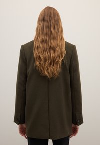 Blazer de lana verde oliva oscuro con una forma estructurada, que presenta mangas largas, una costura central en la parte trasera y puños con botones.