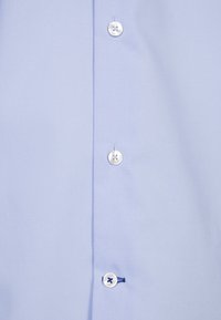 Tissu bleu clair avec trois boutons blancs alignés verticalement sur une patte de chemise, une boutonnière cousue avec du fil bleu foncé.