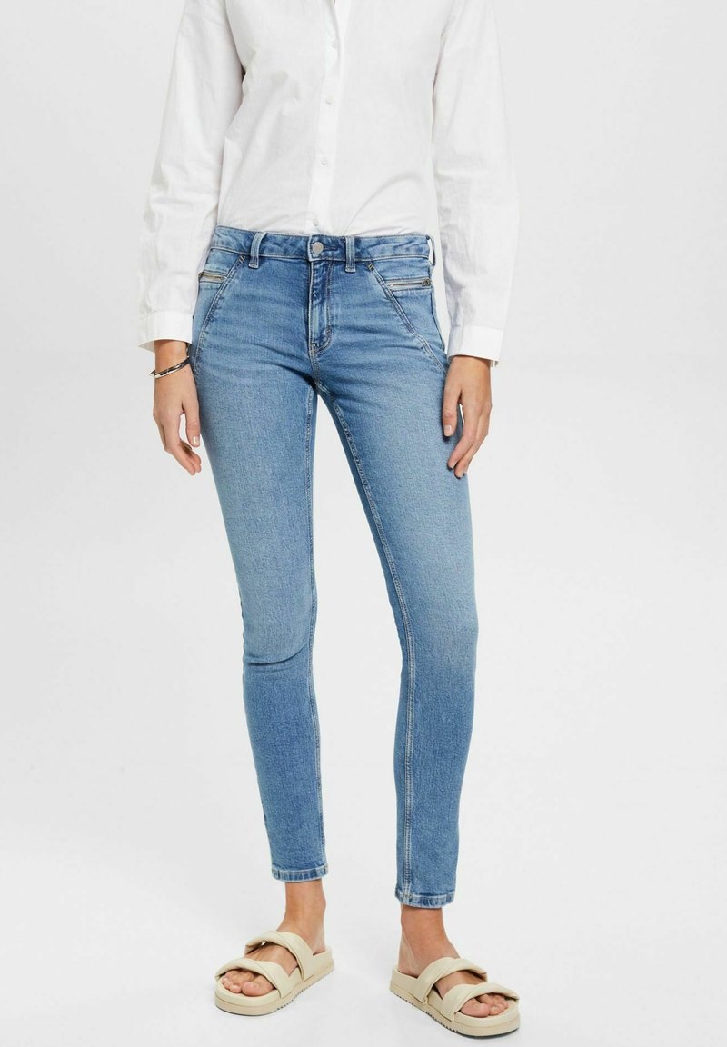 Personne portant un jean skinny bleu clair, une chemise blanche à boutons et des sandales beige à enfiler, debout devant un fond blanc uni.