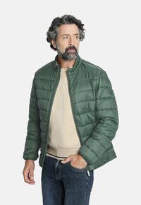 Chaqueta acolchada verde con cuello alzado, cierre frontal y textura acolchada. Llevada sobre una camisa beige y jeans azules.
