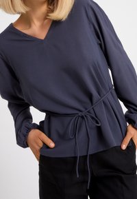 Mörkblå långärmad topp i slätt tyg, med V-ringning och en cinchad midja med snörning, tillsammans med svarta byxor.