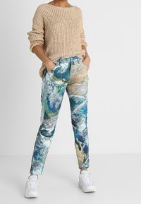 Pletený béžový svetr s širokým výstřihem, kombinovaný s barevnými tištěnými kalhotami s abstraktním designem země v modrých a zelených tónech.