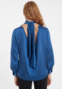 Blaue Satinbluse mit langen Ärmeln, Rückenschlitz und einer Schleife am Hals. Glatte Textur mit einem leicht glänzenden Finish.