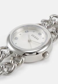 Montre en acier inoxydable argenté avec un cadran rond, un fond blanc, des index en forme de diamant et un bracelet en chaîne épais. Logo de la marque visible.