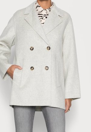 Femme portant un manteau croisé gris clair avec des boutons en écaille de tortue, une chemise à imprimé zèbre, un jean gris, main dans la poche du manteau.