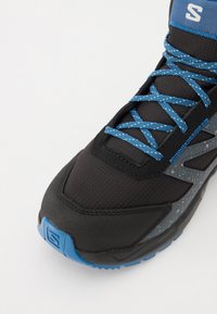 Zapatilla deportiva negra con cordones azul salpicado, acentos grises, tela texturizada y suela de goma con patrón. Logo visible en la lengüeta.