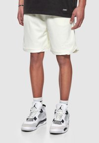 Shorts en maille jaune clair avec une finition texturée, dotés d'un ourlet roulé et d'un logo brodé, associés à des baskets de sport blanches et noires.