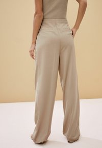 Pantalones de pierna ancha beige con una textura suave, que presentan una cinturilla ajustada y dos bolsillos traseros, situados sobre un fondo liso.