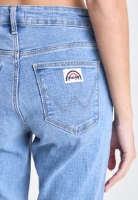 Prikaz izbliza svijetloplave stražnje džepne torbice traperica s logom Wrangler i ruka koja počiva pored kuka.