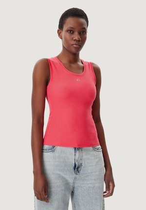 Mujer con una camiseta sin mangas coral de canalé y jeans de mezclilla azul claro, de pie contra un fondo liso y claro.