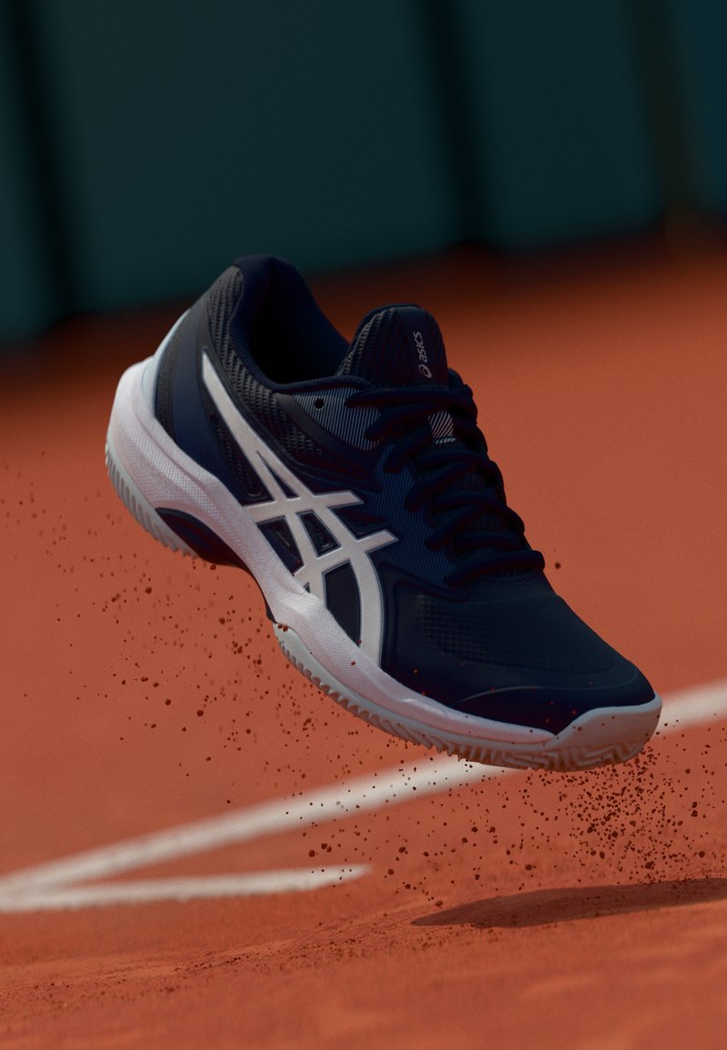 Zapato deportivo de color navy con acentos blancos, parte superior texturizada y suela con patrón, flotando sobre una superficie de arcilla con polvo disperso.