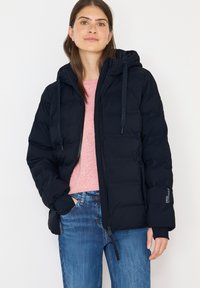 Mørkeblå polstret jakke med hætte, der har elastiske manchetter, frontlynlås og snoretræk. Båret over en lyserød sweater og blå jeans.