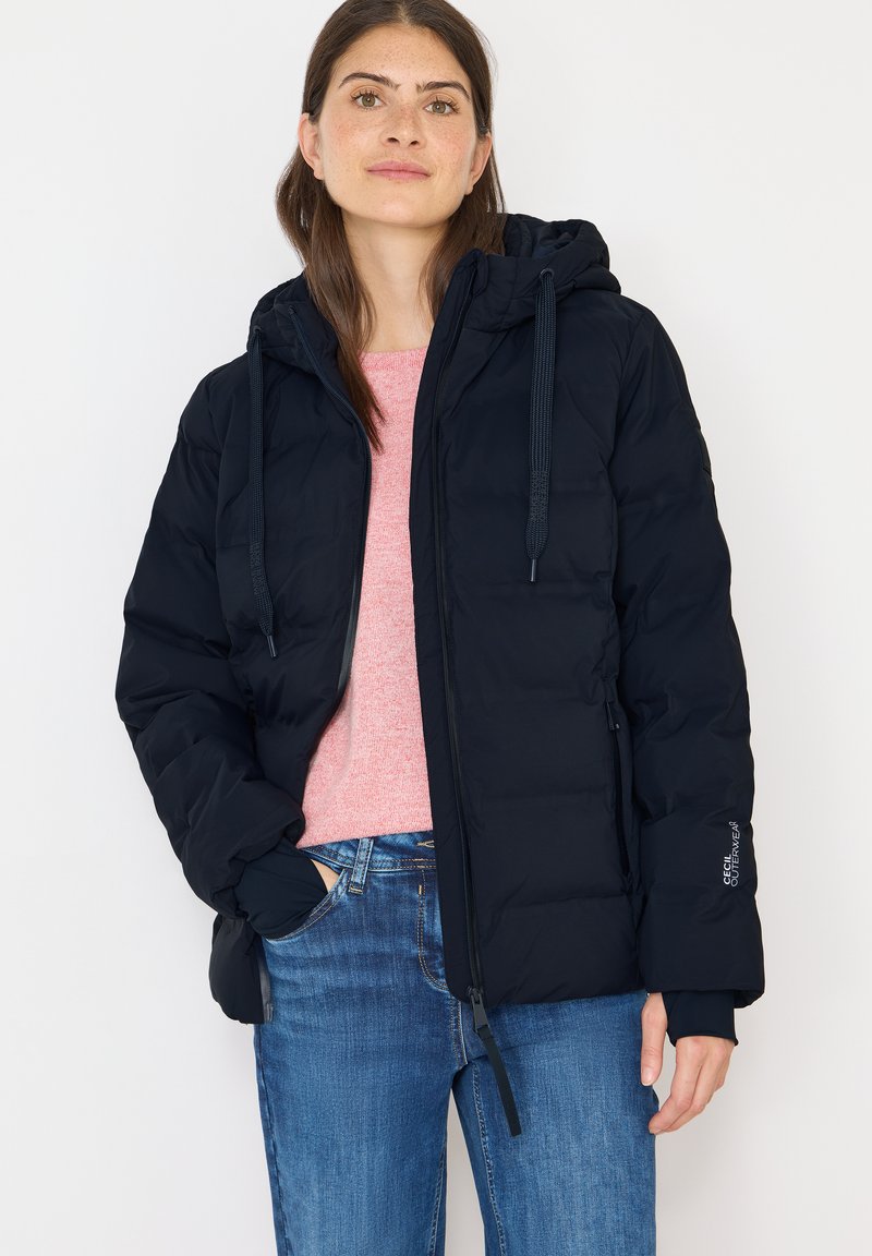 Mørkeblå polstret jakke med hætte, der har elastiske manchetter, frontlynlås og snoretræk. Båret over en lyserød sweater og blå jeans.