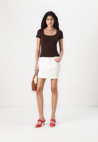 Bruine fitted t-shirt met een vierkante halslijn, gecombineerd met een witte denimrok. Accessoires zijn een bruine handtas en rode hoge hakken.