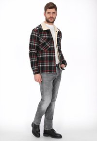 Chaqueta de camisa a cuadros en negro, rojo y gris con un cuello de borrego crema. Cuenta con dos bolsillos en el pecho y mangas ajustadas. Se lleva con vaqueros grises y botas negras.
