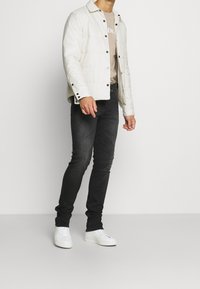 Vadderad off-white jacka med svarta knappar över en beige skjorta, tillsammans med mörk denimjeans och vita sneakers, vilket skapar en lager-på-lager-look.
