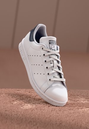 Biały sneaker Adidas Stan Smith z czarną zakładką na pięcie, związanymi sznurowadłami i logo na języku, ustawiony na teksturowanej brązowej powierzchni na rozmytym tle.