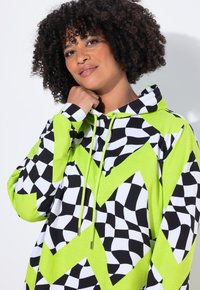 Donna con capelli ricci che indossa un hoodie a scacchi neon verde, nero e bianco, mentre tocca delicatamente il collo, sorridendo dolcemente su uno sfondo semplice.