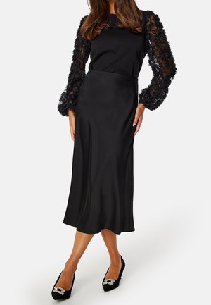 Mujer que lleva una falda midi negra, top negro con mangas abullonadas de encaje floral transparente y tacones negros adornados con punta afilada, de pie sobre fondo blanco.