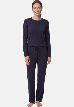 Vrouw staat blootsvoets, draagt een donker top met lange mouwen en bijpassende broek met koord, glimlacht, op een lichte effen achtergrond.