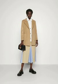 Kamel-farbener, übergroßer Mantel mit Klappentaschen, weißes Hemd mit farbenfrohen Knöpfen und ein beiges Rock, kombiniert mit klobigen schwarzen Schuhen und einer schwarzen Clutch.