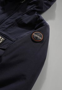 Manica di una giacca blu navy con un patch rotondo nero e arancione del logo Napapijri sulla parte superiore del braccio e una cerniera frontale parzialmente visibile.