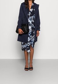 Marinblå trenchcoat, blommig klänning i blått och svart, svart quiltad handväska och svarta öppna tå-klackar. Slät tyg och lager på lager-design.