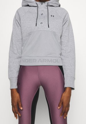 Person iført grå Under Armour hættetrøje med frontlomme og vinrøde og sorte tætsiddende leggings, stående med afslappede armbevægelser.