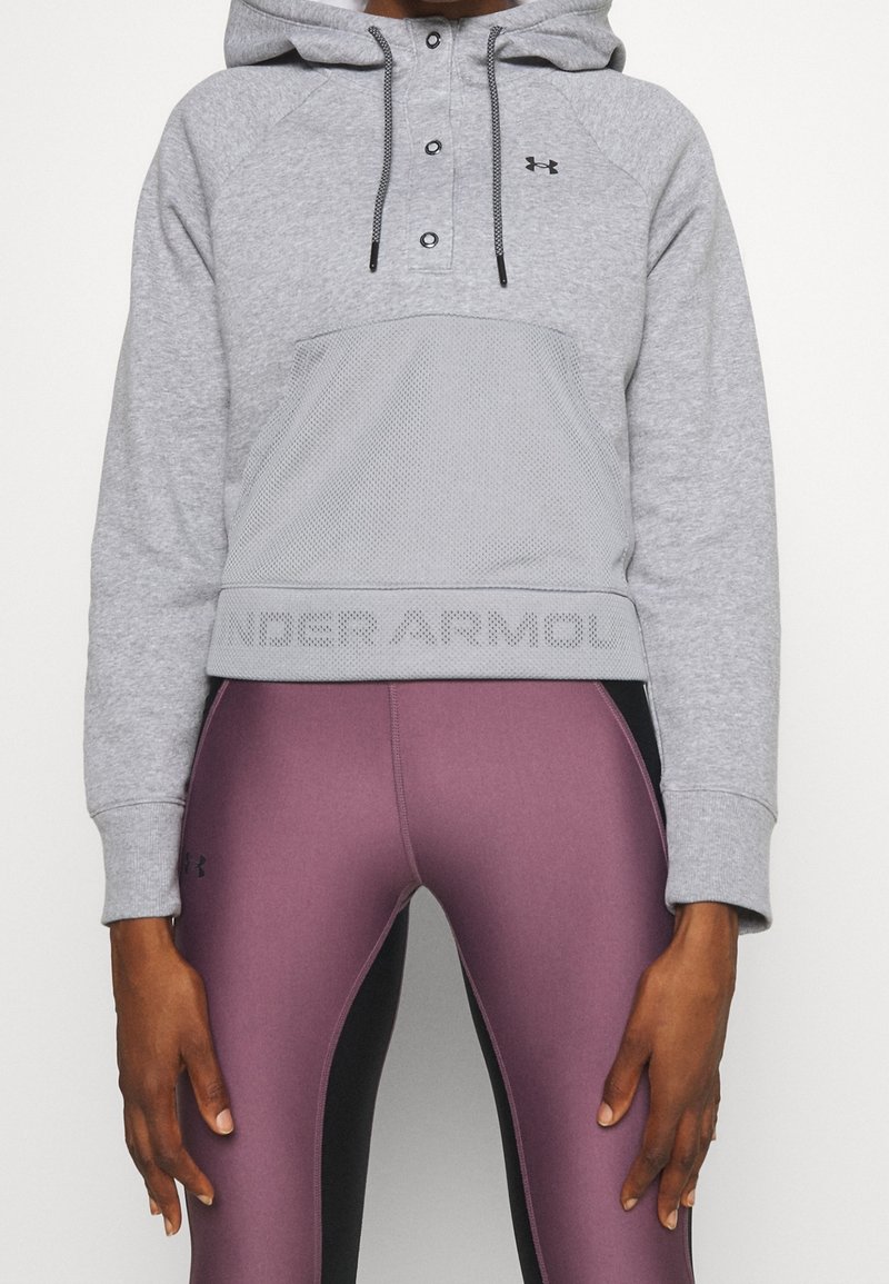 Personne portant un sweat à capuche gris Under Armour avec poche avant et des leggings ajustés bordeaux et noirs, debout, les bras détendus.