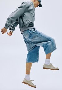 Jachetă bomber gri, pantaloni scurți din denim albastru, cu croială lejeră, și pantofi gri deschis cu tălpi maro. Șosete albe până la glezne vizibile.
