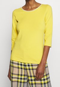 Gelbes Langarmshirt mit rundem Halsausschnitt, versehen mit einem kontrastierenden hellgrauen Besatz. Getragen mit karierten Hosen in Beige, Gelb und Schwarz.