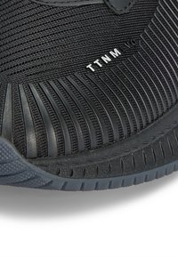 Zbliżenie na czarny, teksturowany but sportowy z karbowaną gumową podeszwą i białym napisem "TTNM" na boku.
