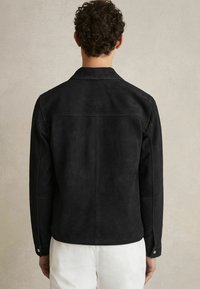 Veste en daim noir avec un col standard, des manches longues et des poignets à pression. Présente une texture lisse et un design épuré et minimaliste.