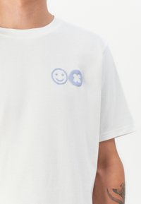 Hvit bomull T-skjorte med lys blå broderte smilende ansikter og blomstermotiver på venstre brystområde. Korte ermer, rund hals.