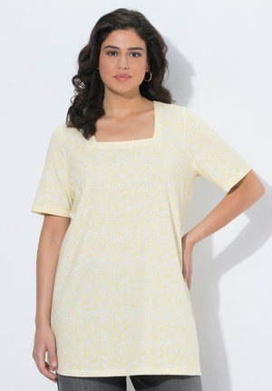 Vrouw met donker golvend haar, gouden oorringen in hoepelvorm en een lichtgeel, vierkant-nek, kortermig topje met patroon, poserend met één hand op haar heup.