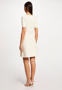 Helles beige Kurzarmkleid mit tailliertem Oberteil, schwingendem Rock und dekorativen Knöpfen entlang der Taille. Glatte Textur, minimalistisches Design.