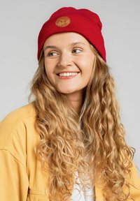 Czerwona dziana czapka z okrągłą skórzaną łatką. Noszona z żółtą koszulką, na tle neutralnym. Częściowo widoczne kręcone włosy.