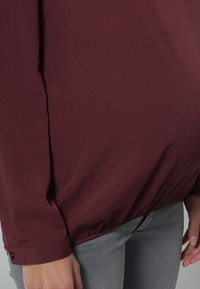 Haut à manches longues bordeaux avec une coupe légèrement ample, ourlet froncé et détails au niveau des poignets, associé à un jean en denim gris. Texture de tissu lisse et léger.