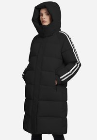 Giacca a vento nera con cappuccio staccabile, motivo a cuciture verticali, tasche laterali e strisce bianche lungo le maniche. Design a lunghezza completa.