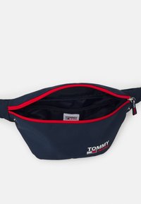 Marinblå midjeväska i slitstarkt material med röd dragkedja, utrustad med en logopatch och ett huvudfack för förvaring.