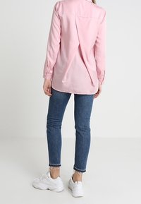 Pink long-sleeve blouse with a relaxed fit, back pleat detail, paired with dark blue jeans and white athletic shoes.