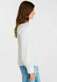 Witte blouse met een getailleerde pasvorm, lange mouwen en een afgeronde kraag, gecombineerd met lichtblauwe spijkerbroek. Eenvoudig en strak design.