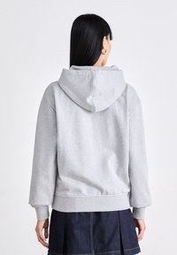 Femme aux longs cheveux noirs portant des boucles d'oreilles créoles dorées, un sweat à capuche gris clair et une jupe plissée foncée, vue de dos sur fond blanc.