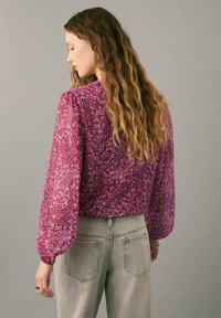Langärmlige, rosa Paisley-Bluse aus transparentem Stoff mit Puffärmeln, kombiniert mit hellgrauen, hoch taillierten Jeans. Strukturierte, florale Muster.